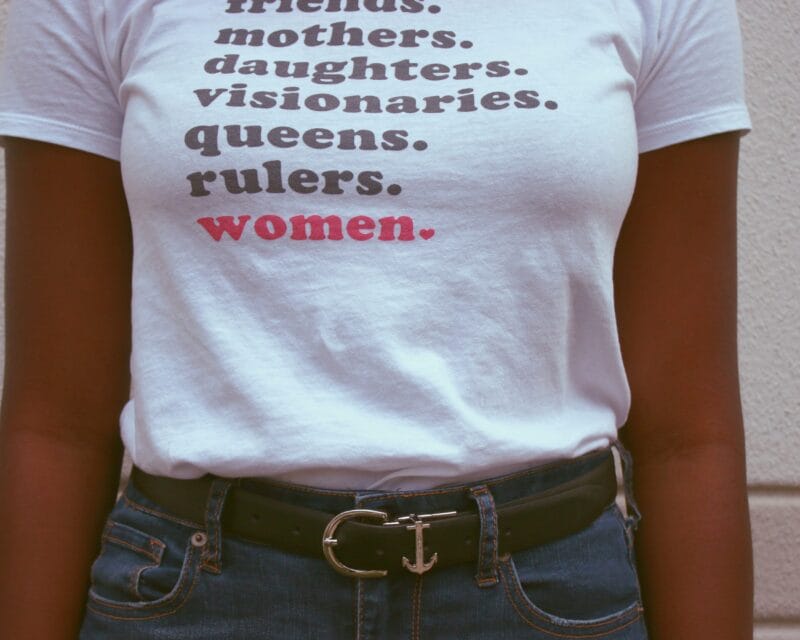 Person wearing shirt reading "Friends. Mothers. Daughters. Visionaries. Queens. Rulers. Women."